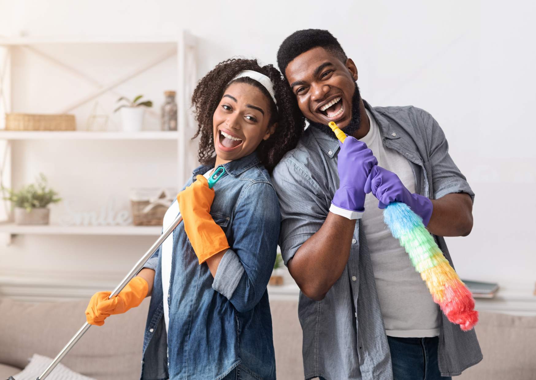 Image shows woman and man playfully posed with cleaning objects. They are in a household setting.